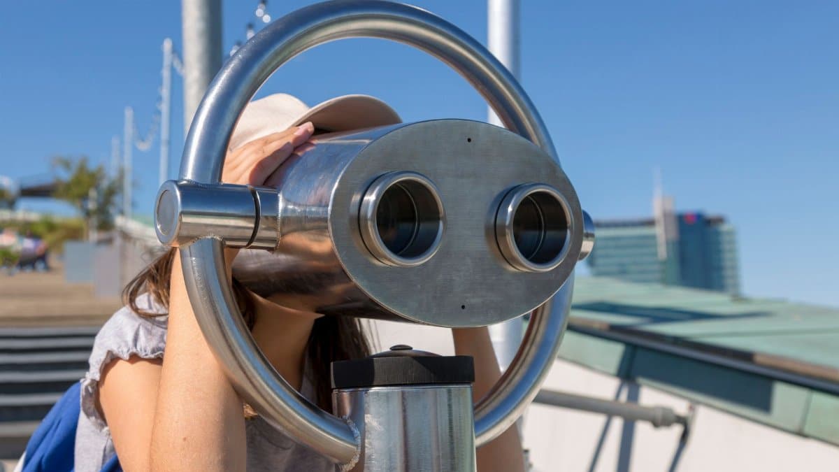 A young woman peering through public binoculars on a sunny day, exploring scenic views.