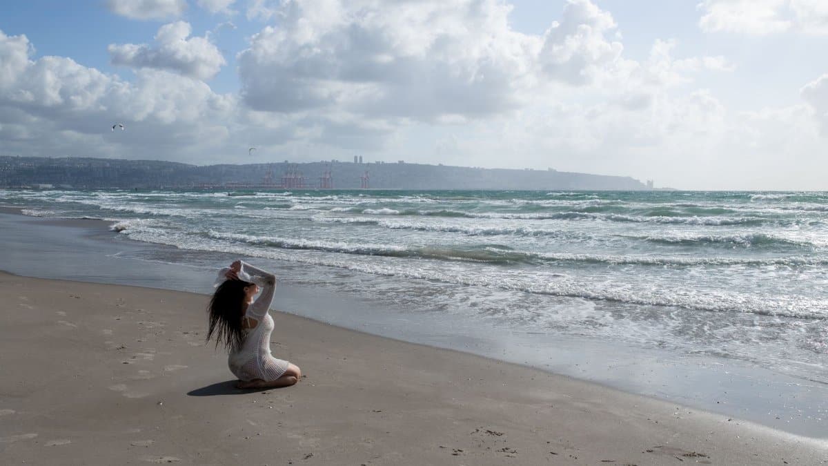 Serene beach scene with a woman meditating, waves crashing and soft clouds in the blue sky.