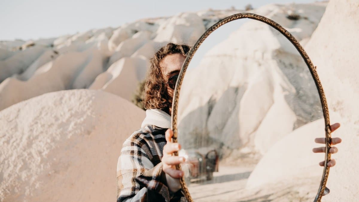 Person in plaid jacket holds mirror reflecting a desert landscape, creating an artistic visual effect.