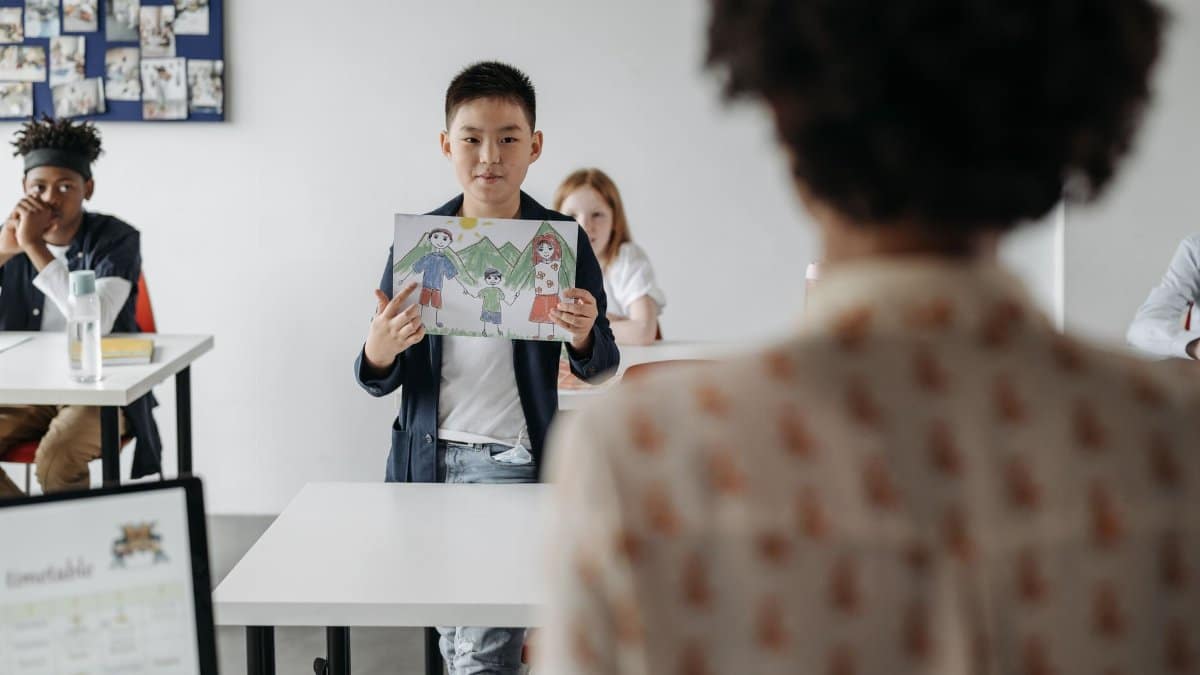 Child holds a drawing in a classroom setting, engaging with peers and teacher.