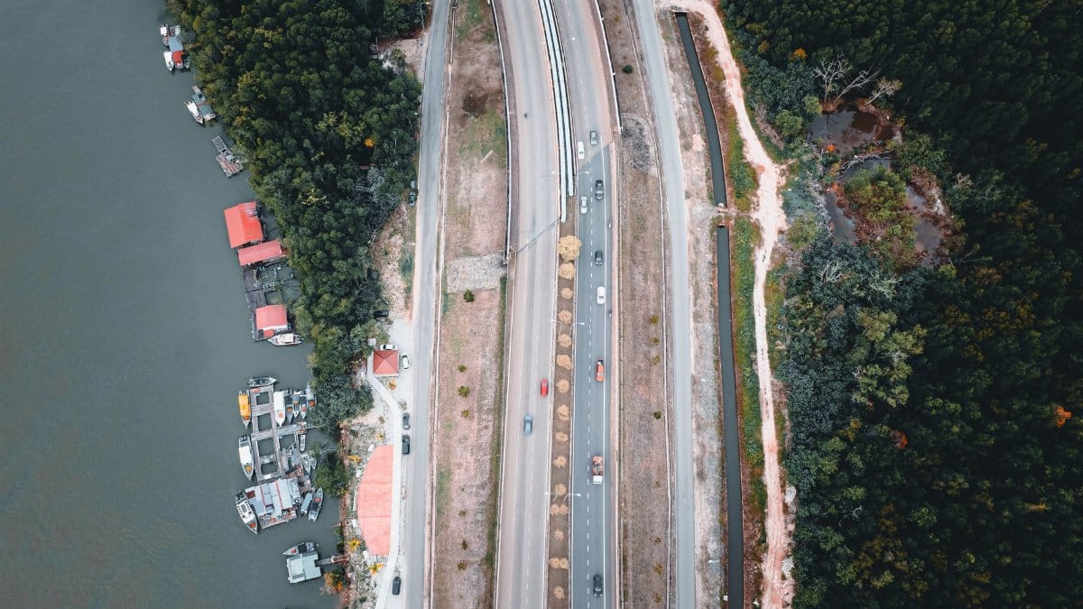 Aerial view of a highway running alongside a river and lush forest, with boats docked nearby.