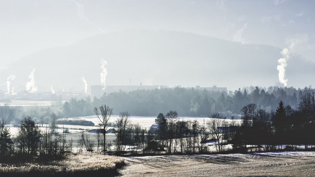 A tranquil winter scene featuring snow-covered fields and distant industrial factories emitting smoke.