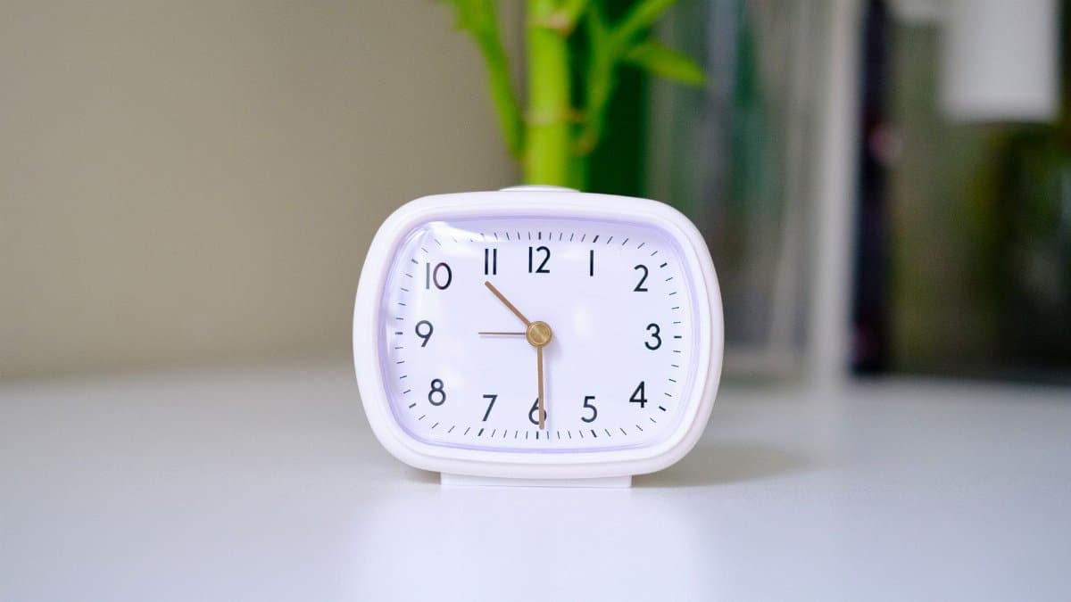 A minimalist white analogue clock placed on a clean desk with a blurred plant in the background.