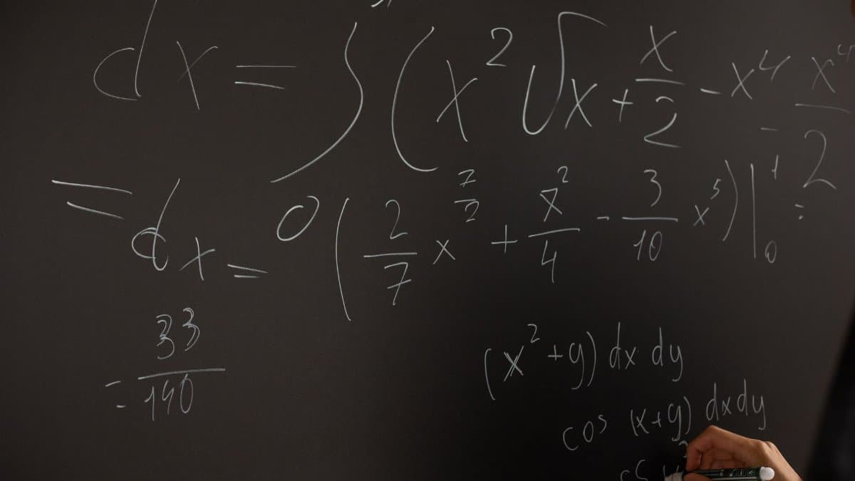 Close-up of hand writing complex math equations on a chalkboard in a classroom setting.