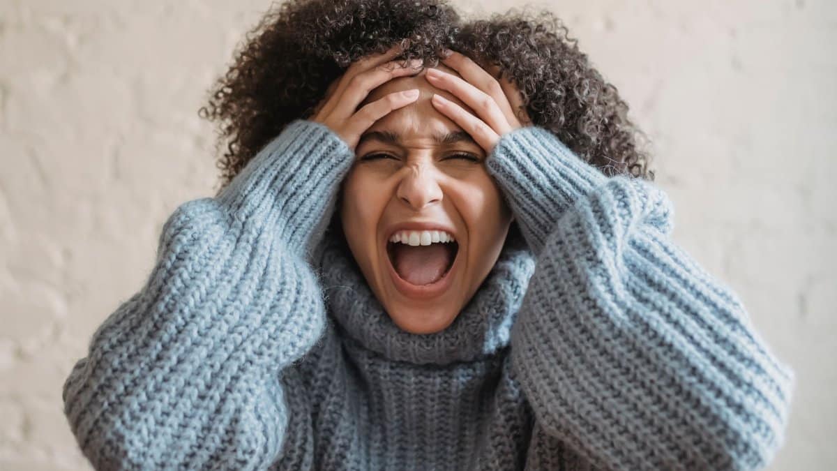 Angry ethnic female with closed eyes in warm wear touching head while shouting in light room near white wall at home