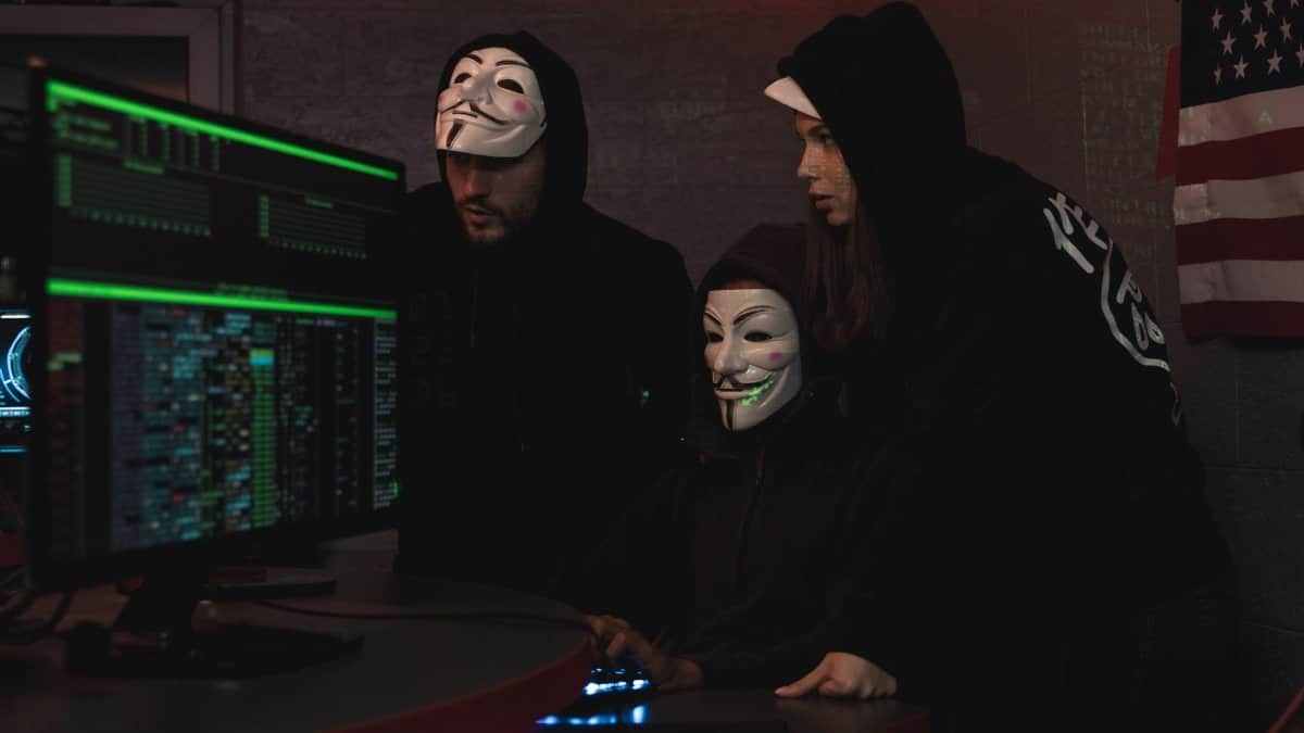 Masked hackers in hoodies working on a system in a dimly lit room, representing cyber security threats.