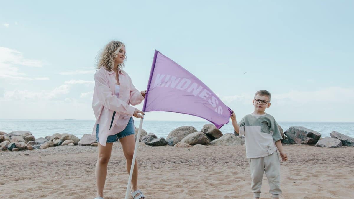 A mother and son hold a kindness flag on a sunny beach, promoting positivity.