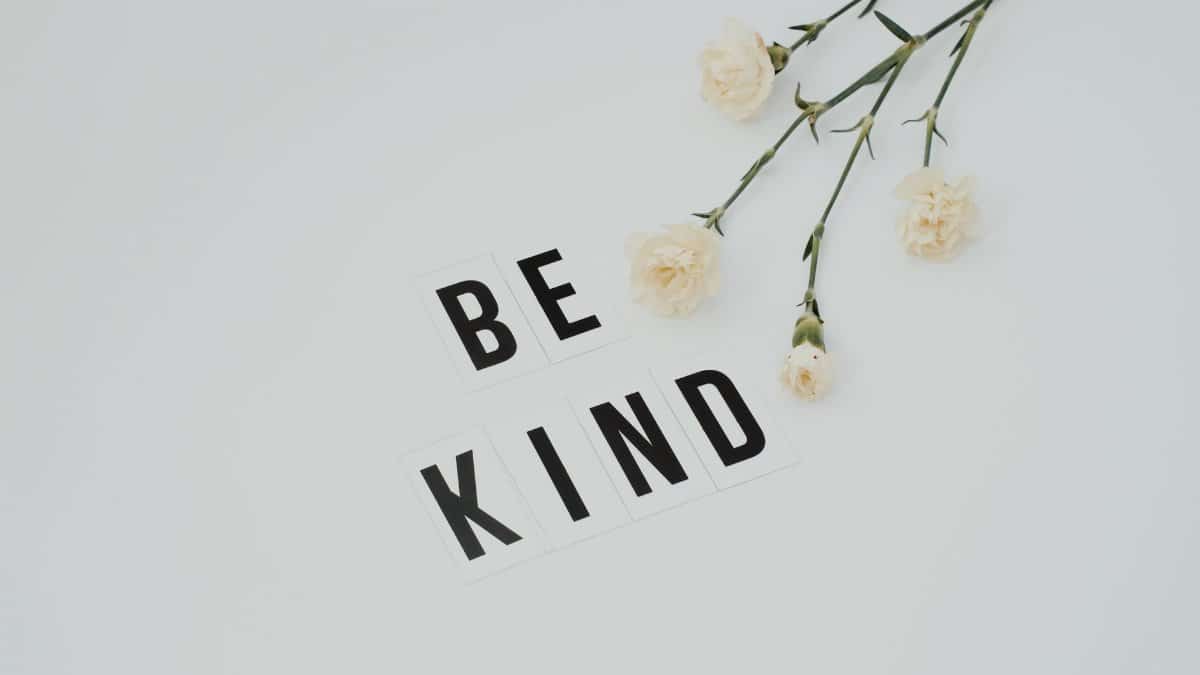 Minimalist image of 'Be Kind' text with white flowers on a light background, promoting kindness.