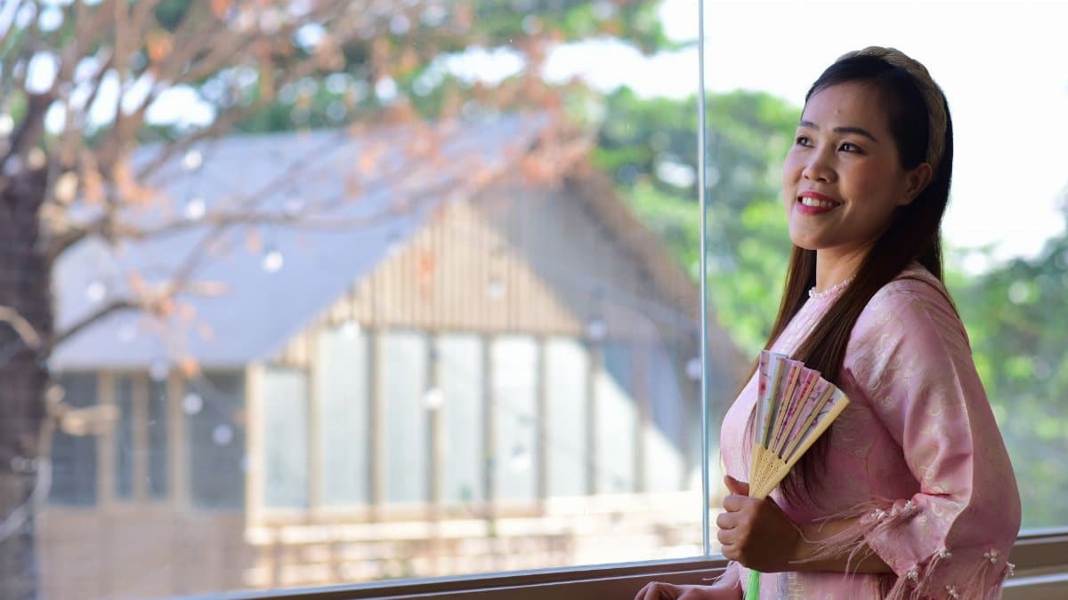 Smiling woman in traditional attire holding a fan by the window with soft light.