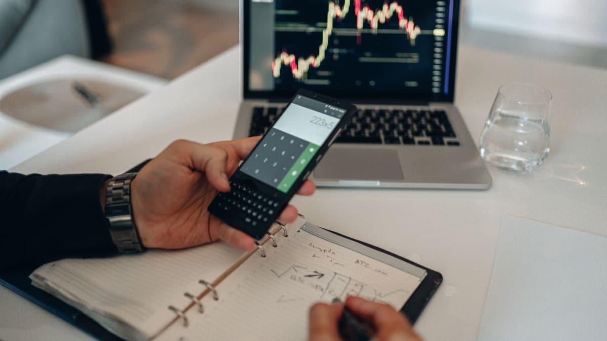 Close-up of a person using a calculator and laptop with stock charts for financial analysis.