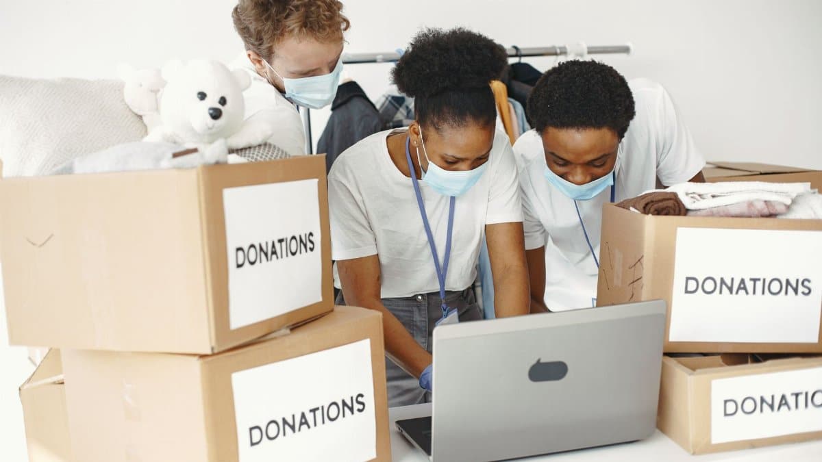 Group of volunteers wearing masks sorting donations with a laptop, promoting diversity and teamwork.