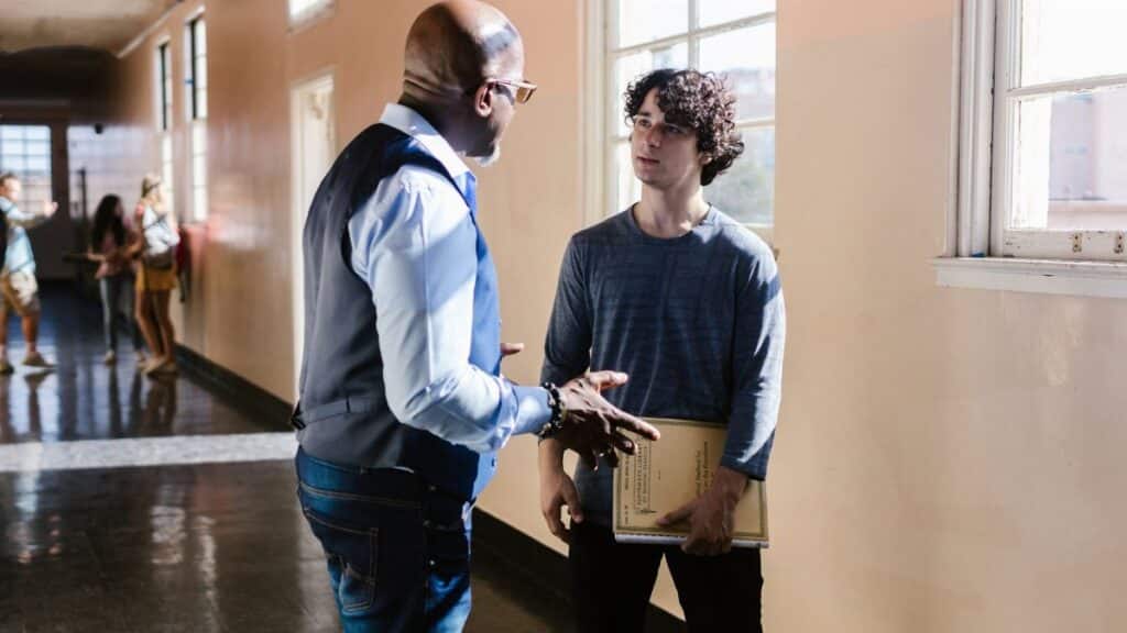 A high school teacher and student having a conversation in a brightly lit hallway.