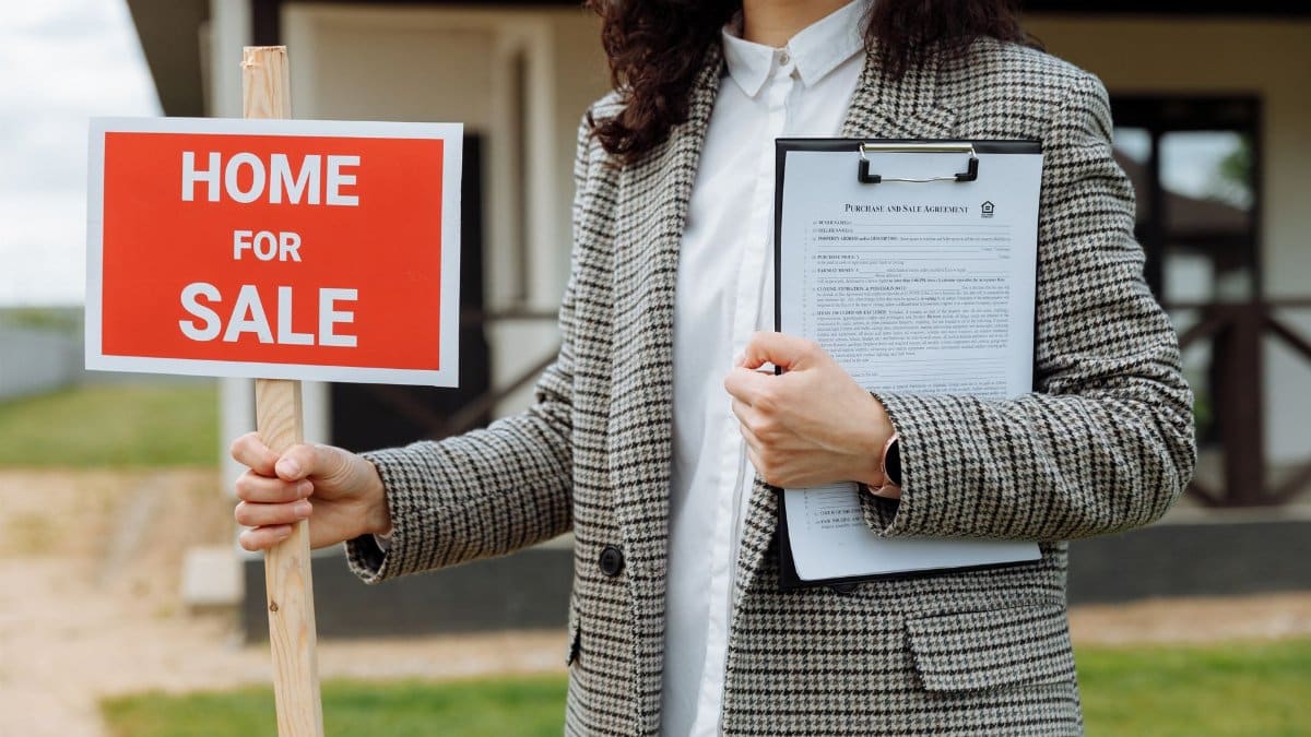 A real estate agent holding a home for sale sign and clipboard outside a property.