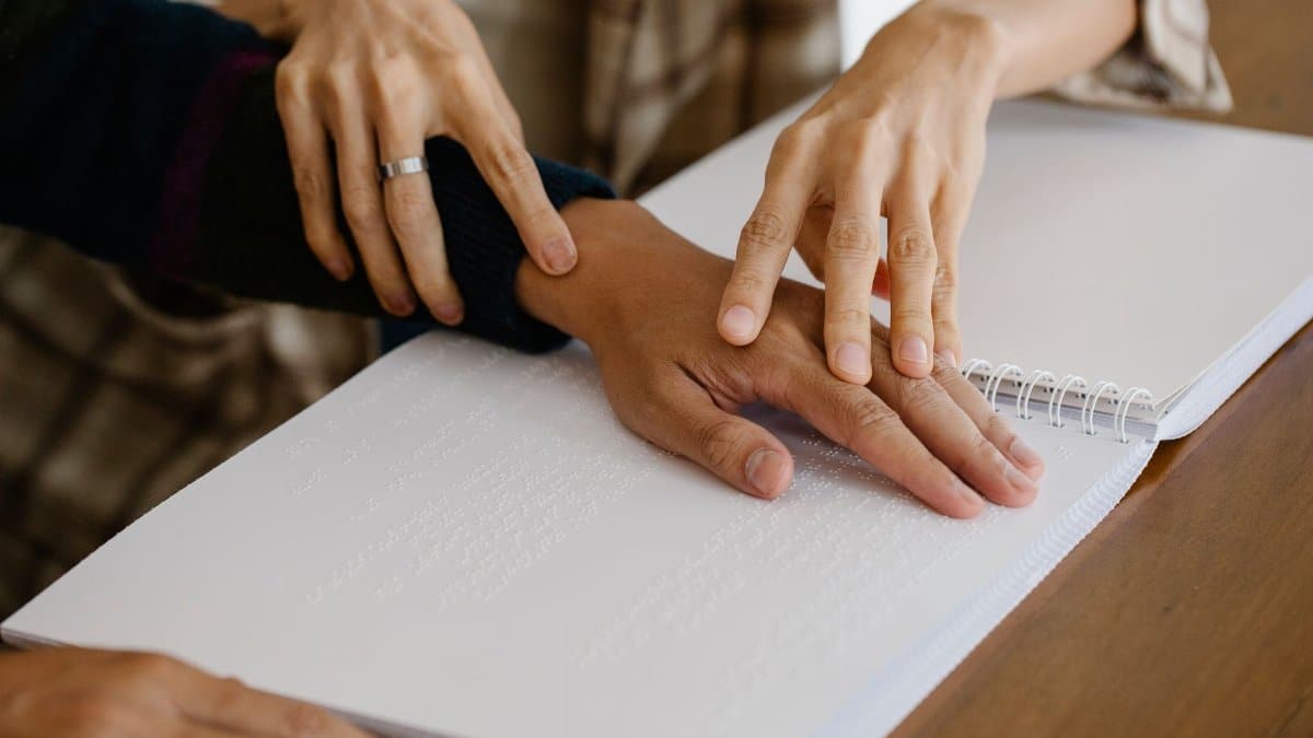 Crop anonymous caregiver guiding hand of blind patient reading information in copybook with braille text