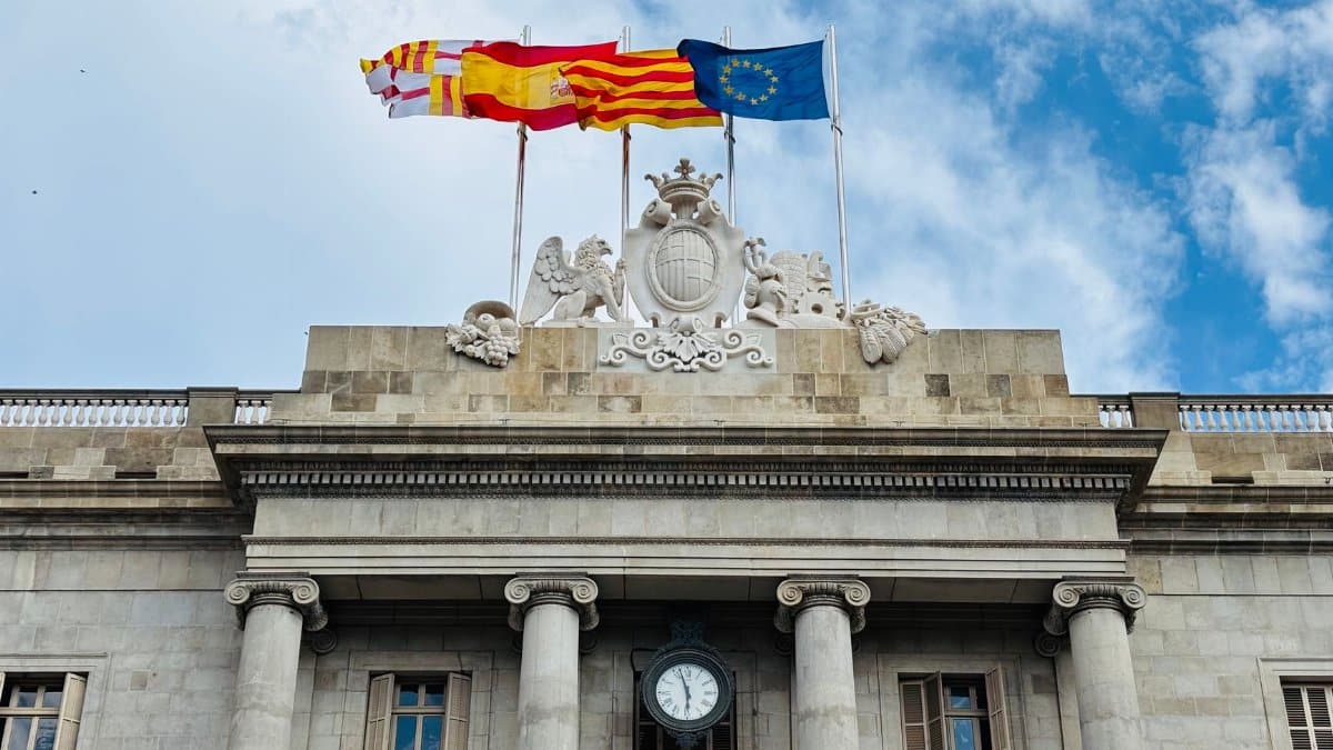 Stone building facade with European, Spanish, and regional flags under blue sky.