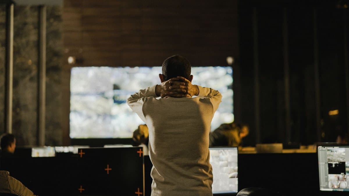 A man stands thoughtfully in a dimly lit control room with various screens.