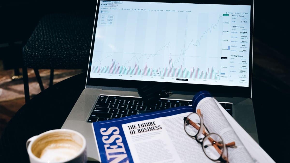 A modern business workspace featuring a laptop showing stock data next to a business magazine and coffee.