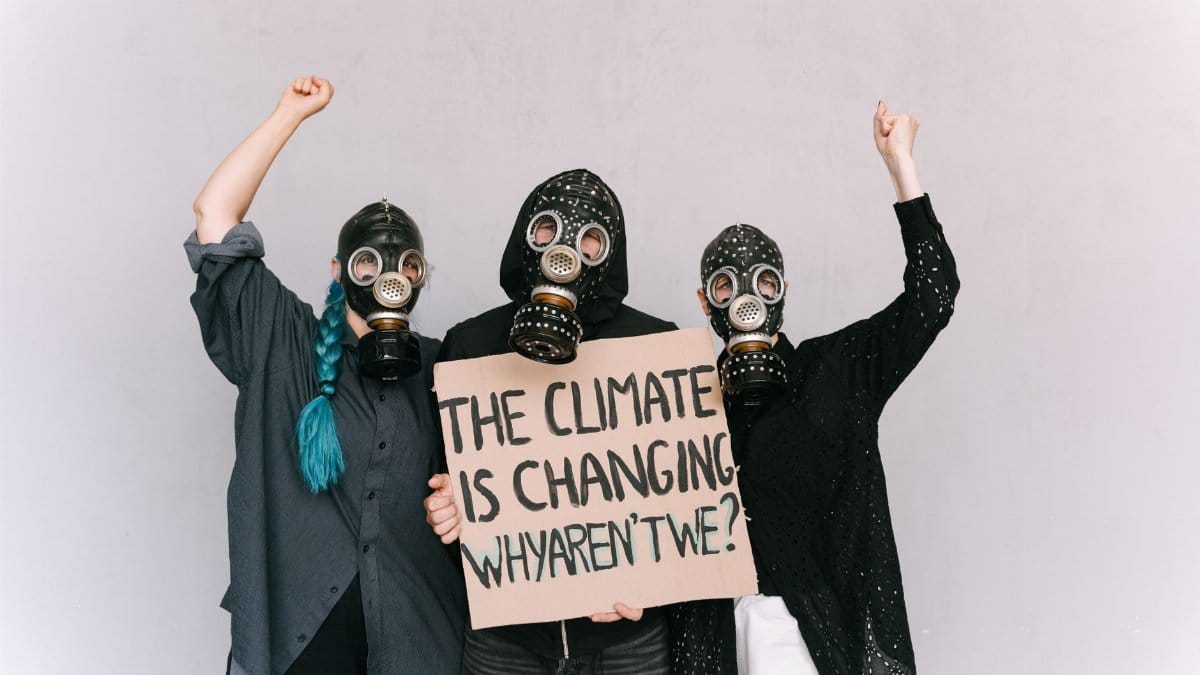 Three activists wearing gas masks hold a climate change protest placard indoors.