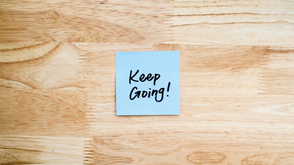 A blue sticky note with 'Keep Going!' message on wood background.