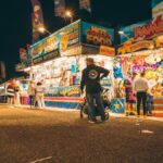 A lively night fair with illuminated stalls, games, and blurred motion of people.