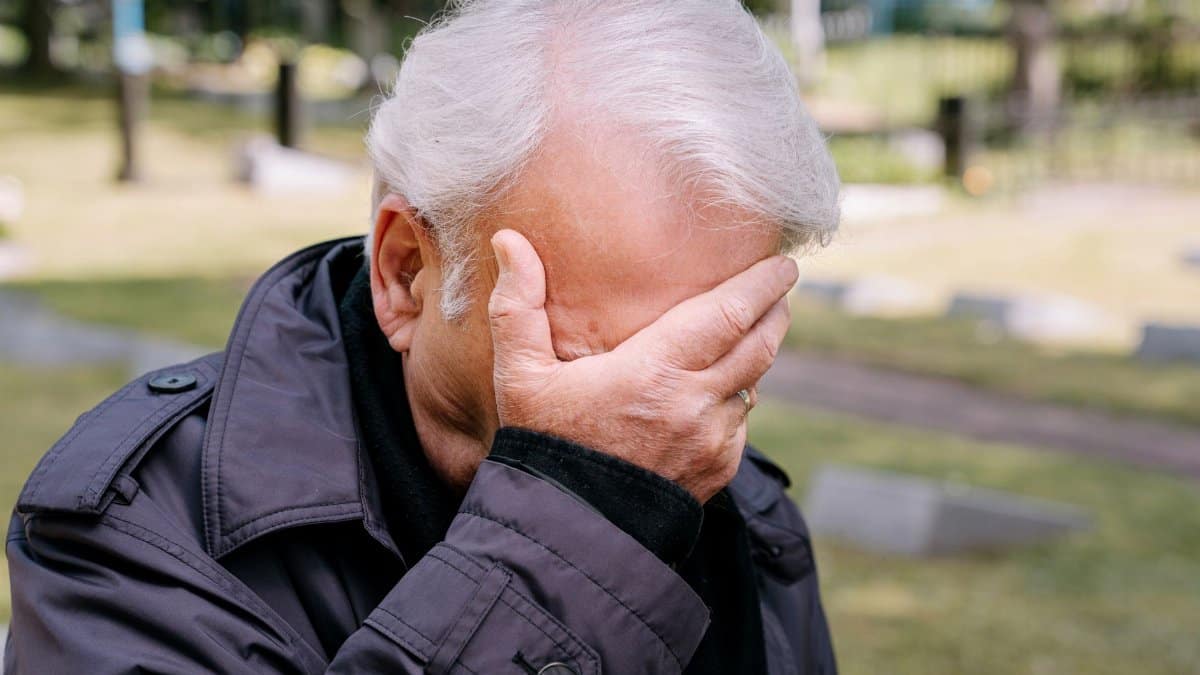 Senior adult man in grief, covering face with hand, in a cemetery setting.