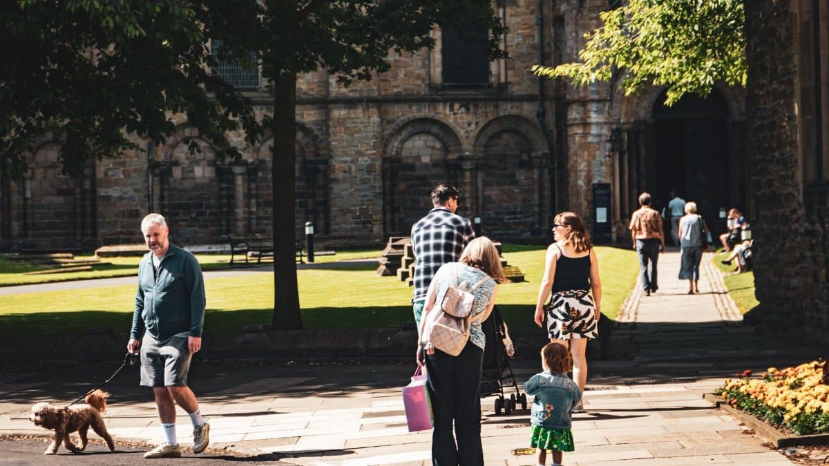 A diverse group strolling through the grounds of historic Durham Cathedral on a sunny day.