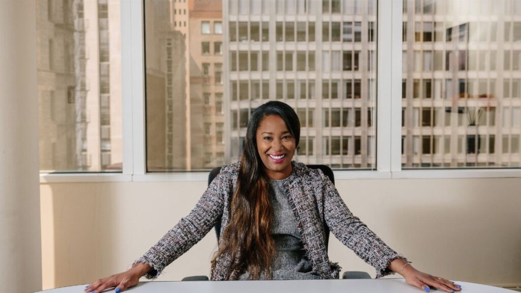 Smiling businesswoman in professional attire seated in a bright office interior with city view.