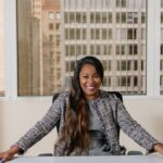 Smiling businesswoman in professional attire seated in a bright office interior with city view.