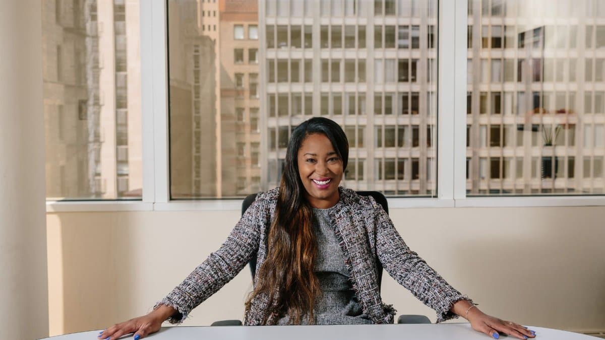 Smiling businesswoman in professional attire seated in a bright office interior with city view.