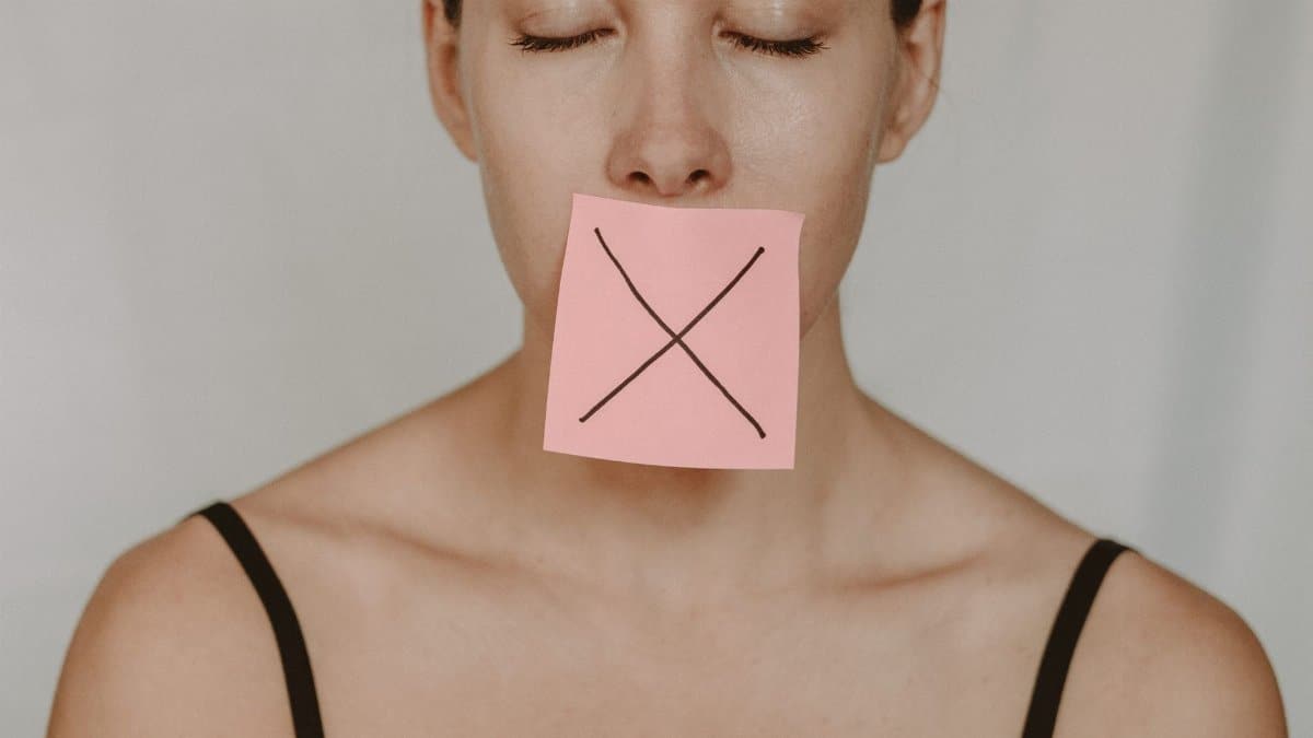 Young woman in black lingerie with closed eyes showing pink sticker with black cross against violence and abuse on lips on white background in light studio