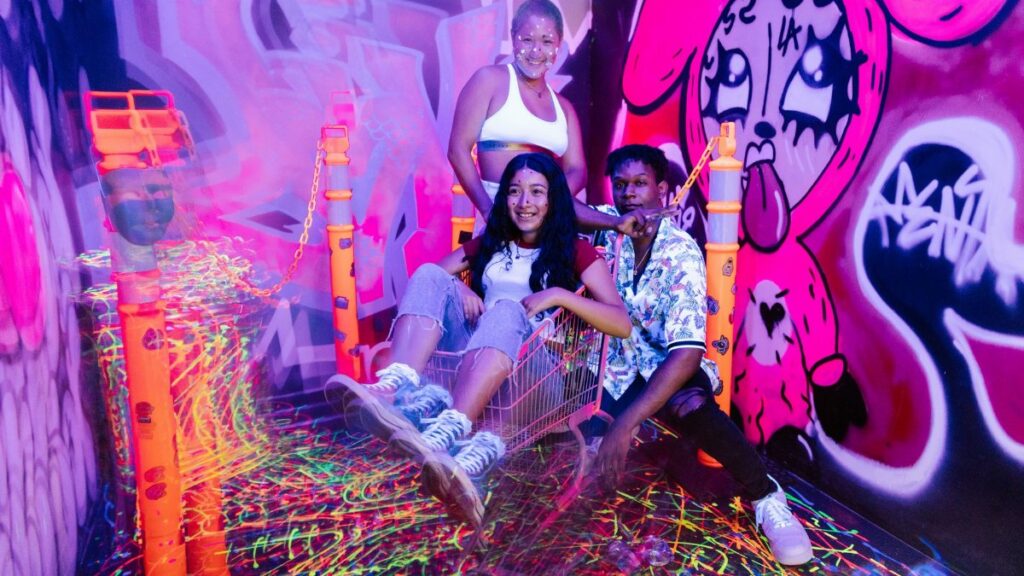 Vibrant neon-lit indoor party scene with friends enjoying a playful shopping cart ride amidst graffiti art.