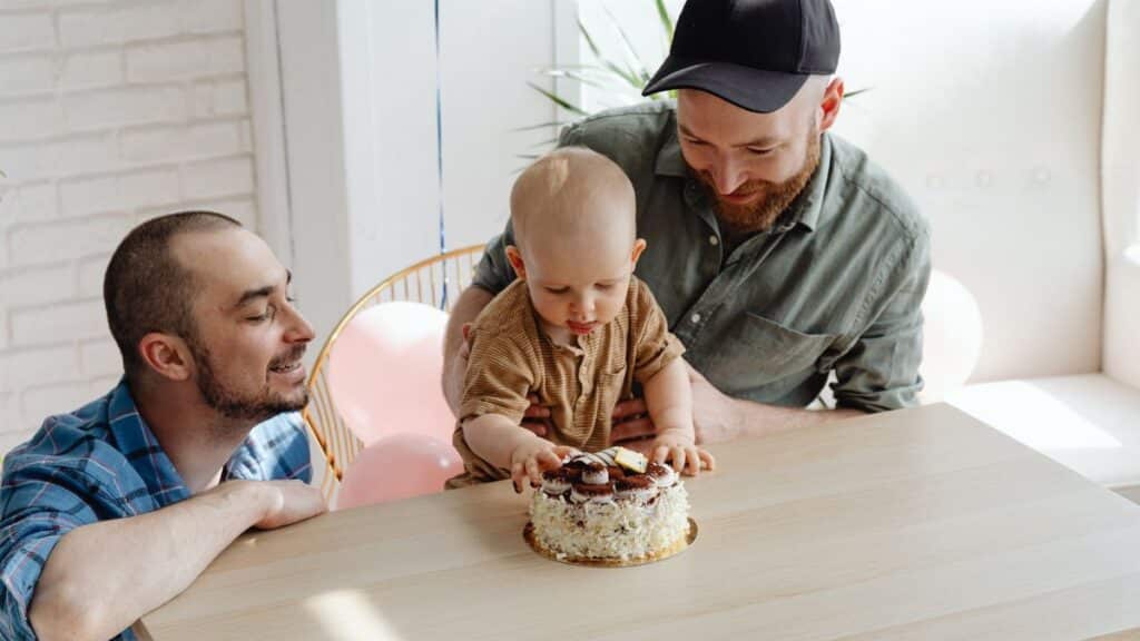 Two fathers with their baby celebrating a birthday with a cake indoors.