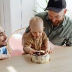 Two fathers with their baby celebrating a birthday with a cake indoors.
