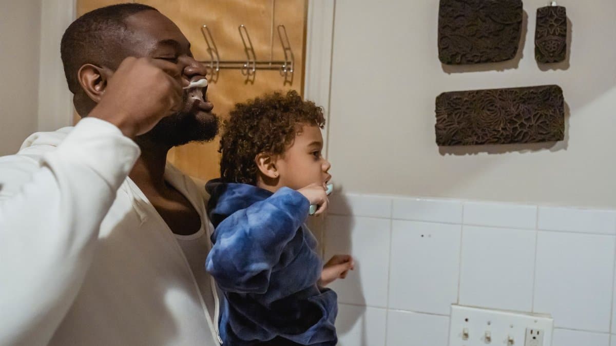 Side view of serious African American father with son in hands brushing teeth while standing in bathroom during daily routine