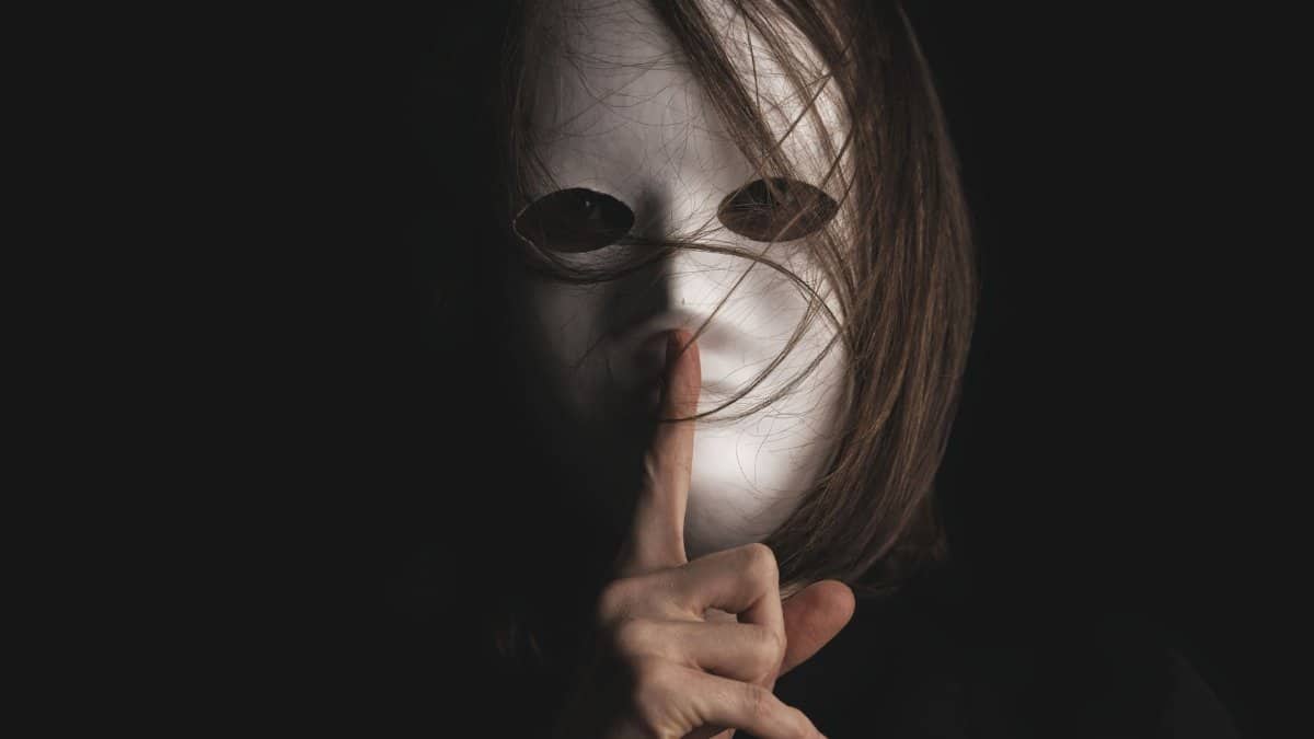 A person with a white mask makes a silence gesture, conveying mystery and secrecy.