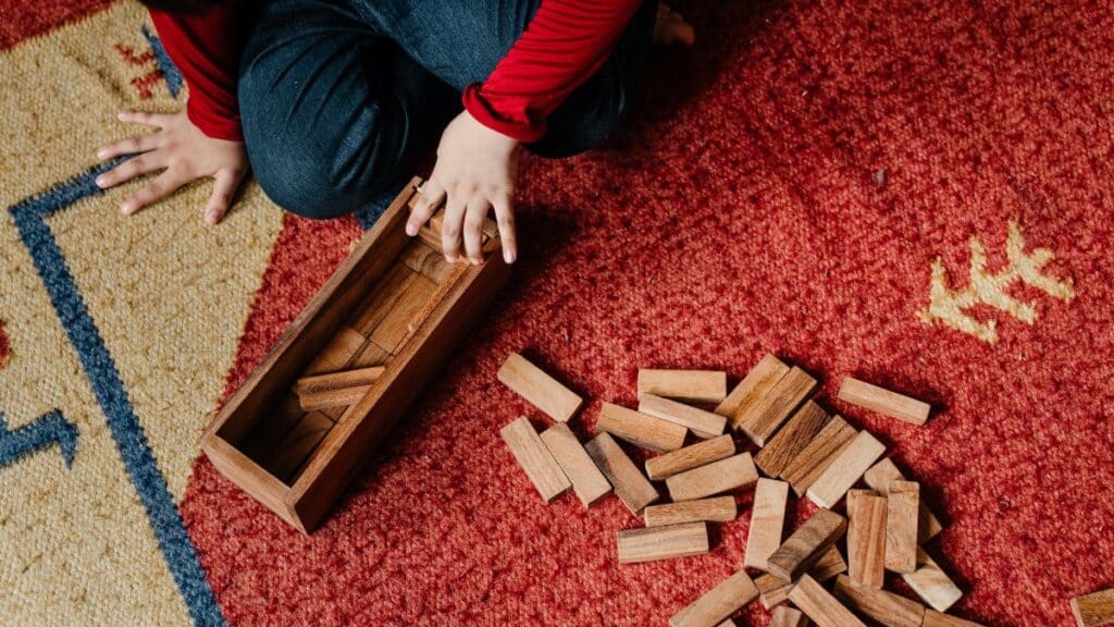 From above crop kid in casual clothes sitting on floor carpet while playing in jenga tower game with wooden blocks improving mental skills and accuracy
