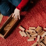 From above crop kid in casual clothes sitting on floor carpet while playing in jenga tower game with wooden blocks improving mental skills and accuracy