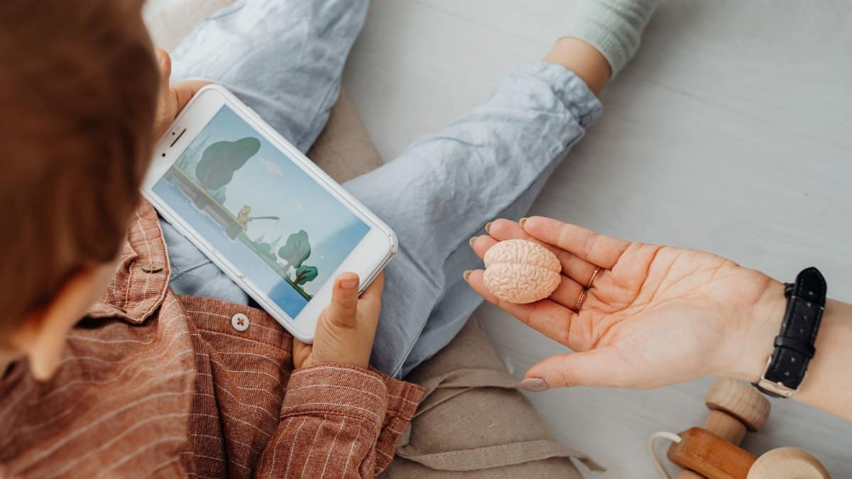 Young child engrossed in a smartphone video while holding a brain model indoors.