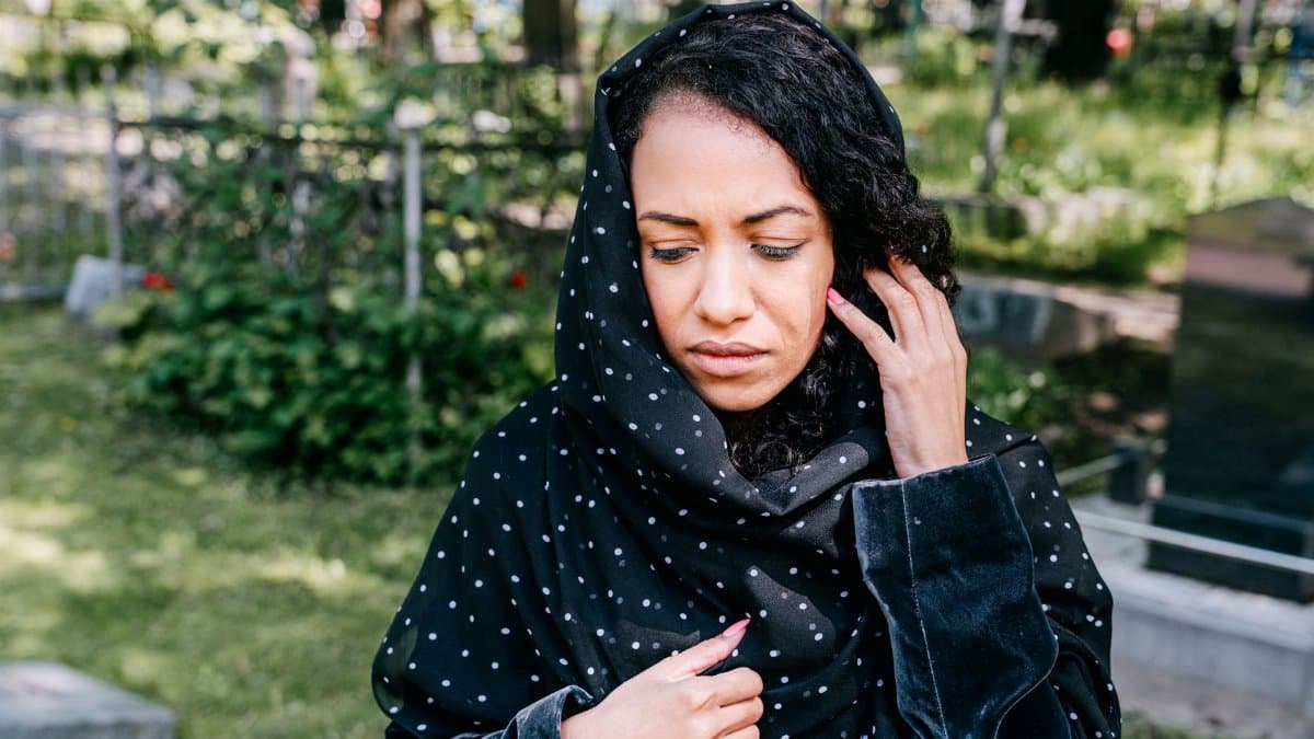 A woman in a cemetery expressing grief, wearing a polka dot headscarf, outdoors.
