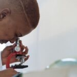 Side view of young African American doctor in uniform examining samples through microscope while working in modern clinic at daytime
