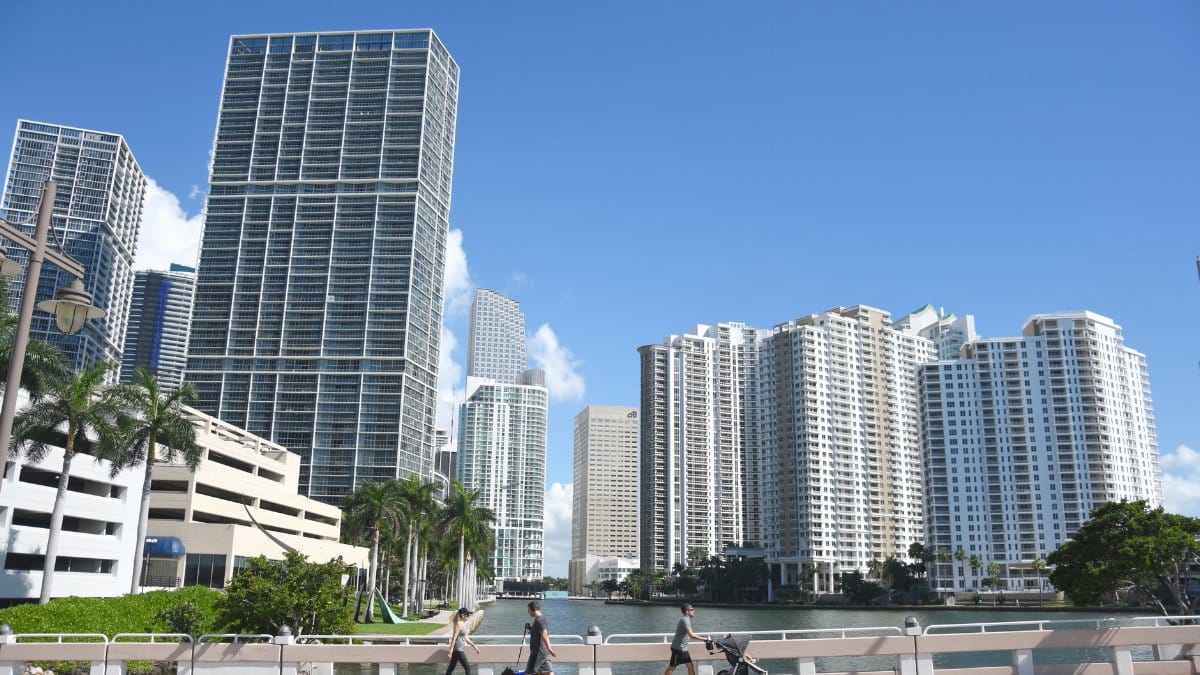 Skyline of modern skyscrapers in downtown Miami under a clear blue sky with pedestrians on a bridge.