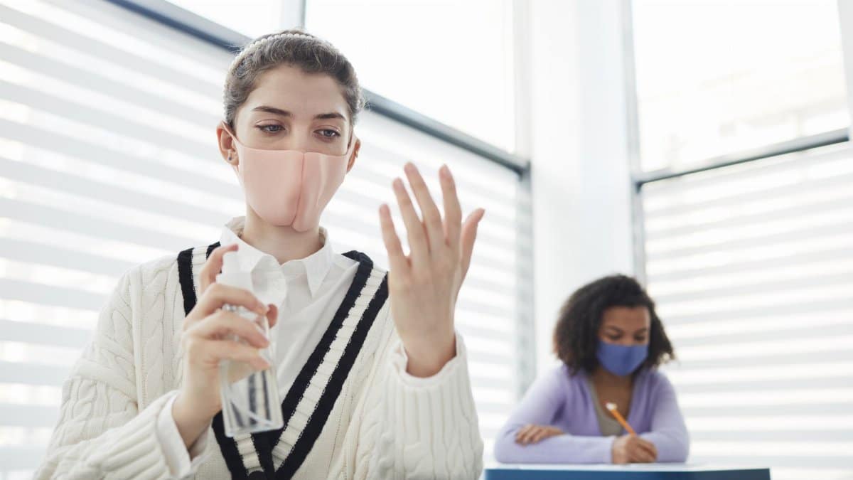 A woman wearing a face mask sanitizes her hands while another sits at a desk in a classroom setting.