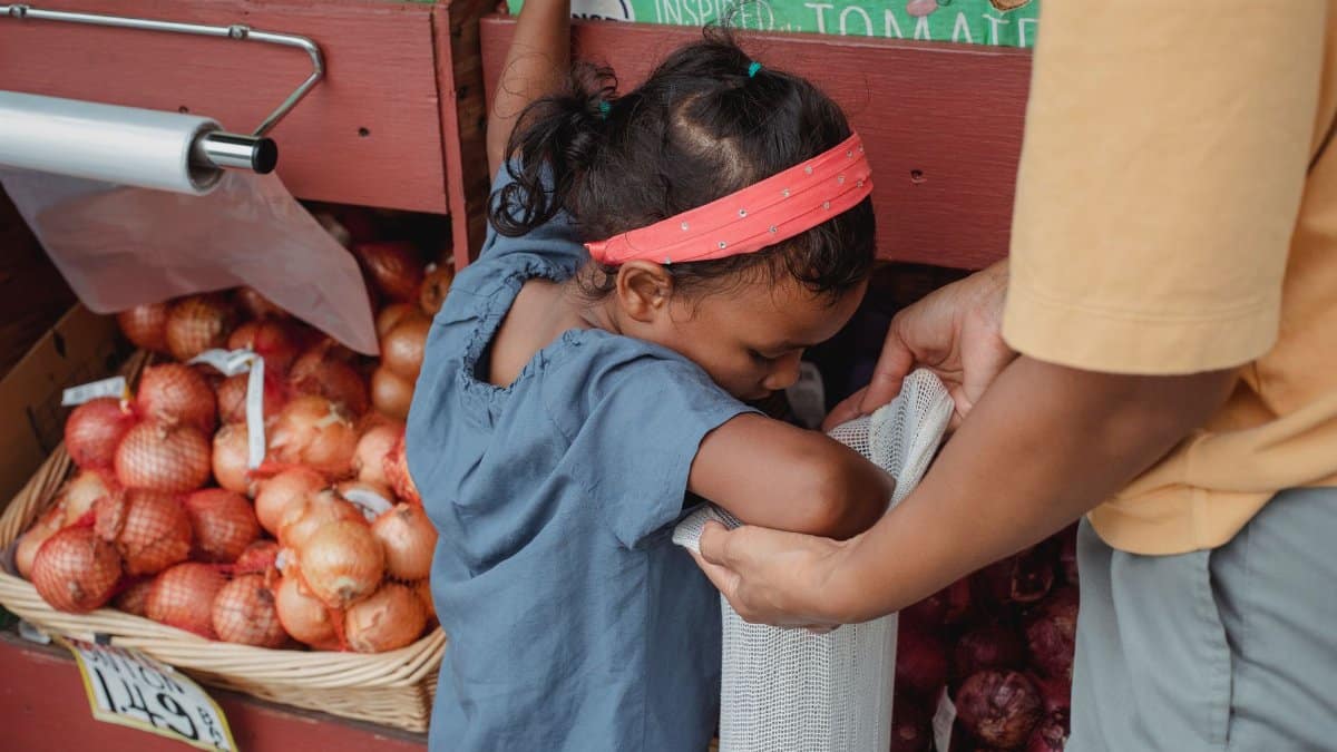 Asian girl and mother putting vegetables in bag while doing groceries in local street market and standing near stall