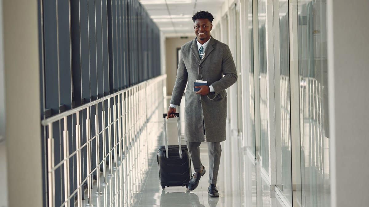 Full length of happy African American male in stylish classy gray clothes with suitcase and passport walking in contemporary airport terminal