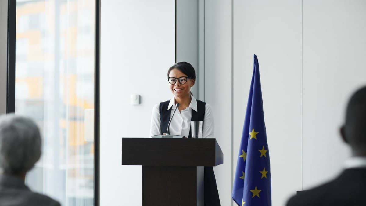 A woman giving a speech at a podium with an EU flag in a conference setting.