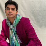 Young man posing confidently in a pink suit with floral scarf, set against a neutral backdrop.