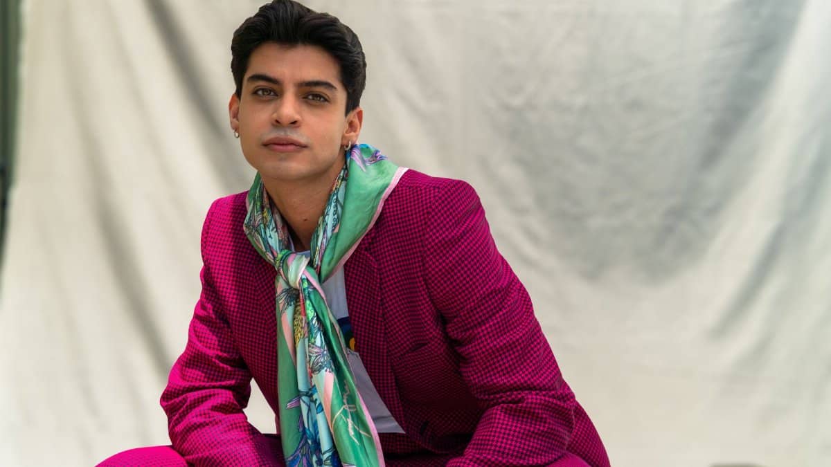 Young man posing confidently in a pink suit with floral scarf, set against a neutral backdrop.