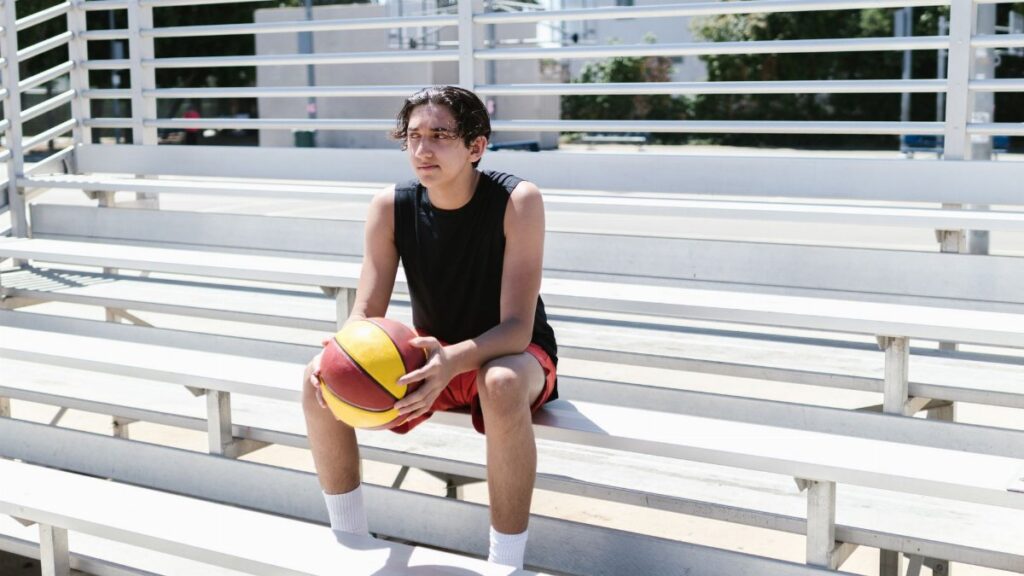 Teen basketball player relaxing on outdoor bleachers during the day.