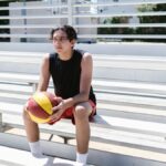 Teen basketball player relaxing on outdoor bleachers during the day.