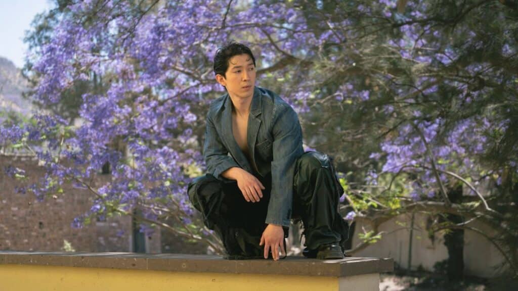 Young man in stylish attire poses outdoors with vibrant jacaranda tree backdrop.